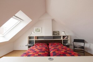 A bedroom with a sloped ceiling, a bed with a checkered bedspread, a desk with a chair, and a shelf with books and a lamp. - Apartment 17, Südstrasse 1 (Norderney)
