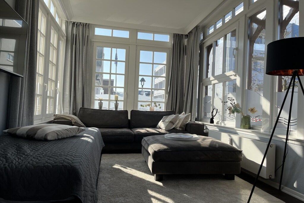 A modern bedroom with a large bed, a sofa, and an ottoman. There are multiple windows providing natural light and a view of the outside.