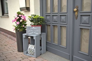 A door with a plaque, a potted plant with pink flowers, and a white decorative item on a small table. - Holiday apartment with 1 bedroom (Borken)