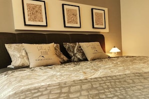 Ein Bett mit gemusterten Betttextilien, drei gerahmte Bilder an der Wand und ein Nachttisch mit einer Lampe.