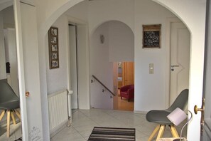 Un couloir avec un escalier menant à une pièce, une porte sur laquelle est accroché un tableau noir servant de panneau, et une chaise avec un coussin.
