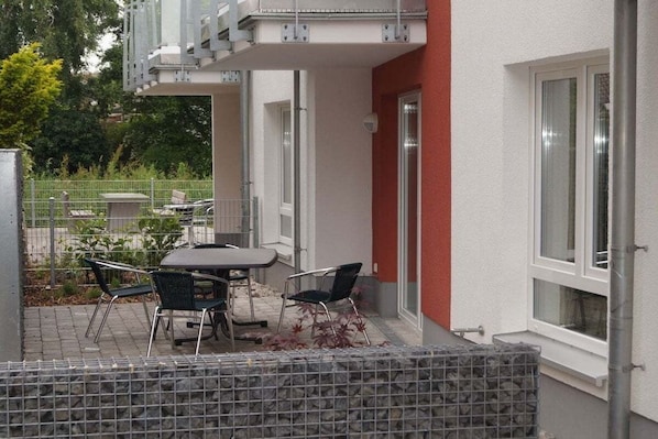Um pátio externo com mesa e cadeiras, um muro de pedra e um edifício com fachada vermelha e grandes janelas.