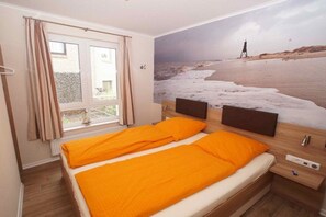 Ein Schlafzimmer mit Bett, einem Fenster mit Vorhängen, einem an der Wand befestigten Fernseher und einem Strandbild.