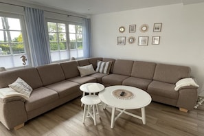 A living room with a sectional sofa, two white round tables, and a large window with curtains. - Holiday home with 1 bedroom (Timmendorfer Strand)