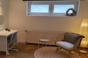 A room with a white desk, a wicker chair with a cushion, a small round table, a radiator, and a window with a wreath. - Holiday home with 1 bedroom (Timmendorfer Strand)
