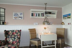Ein gemütliches Zimmer mit einem weißen Tisch und zwei Korbstühlen, einem Bücherregal mit Büchern, einem gerahmten Bild an der Wand und einer kleinen Ablage mit Dekorationen.