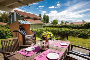 An outdoor dining area with a table set for two, surrounded by wooden chairs, a wicker chair, and a small grill. - Captain's House Type F (Neustadt\/Holstein - OT Pelzerhaken)