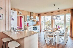 A modern kitchen with a central island, a dining area with white chairs, and large windows. - Captain's House Type F (Neustadt\/Holstein - OT Pelzerhaken)