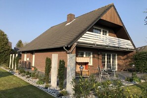 Ein zweigeschossiges Haus mit Satteldach, einer Veranda mit Bank und einem kleinen Gartenbereich mit Pflanzen und einem Weg.