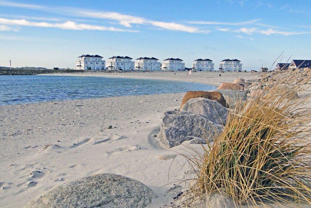 A sandy beach with a row of white buildings with dark roofs, a rocky shoreline, and some dry grass.