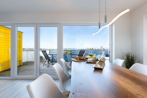 A modern dining area with a wooden table, white chairs, and a view of a yellow building and a body of water through large windows. - Holiday apartment with 2 bedrooms (Kappeln)