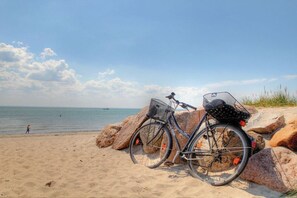 Ein Fahrrad mit Korb und Gepäckträger, abgestellt an einem sandigen Strand mit großen Steinen und dem Meer im Hintergrund.
