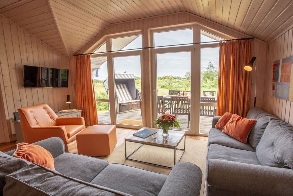 A cozy living room with a sofa, armchair, and coffee table, large windows with curtains, and a view of the outdoors.
