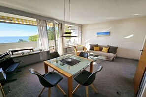 A modern living room with a glass dining table, black chairs, a sofa with cushions, and a view of the sea through large windows. - Holiday apartment with 2 bedrooms (Sierksdorf)