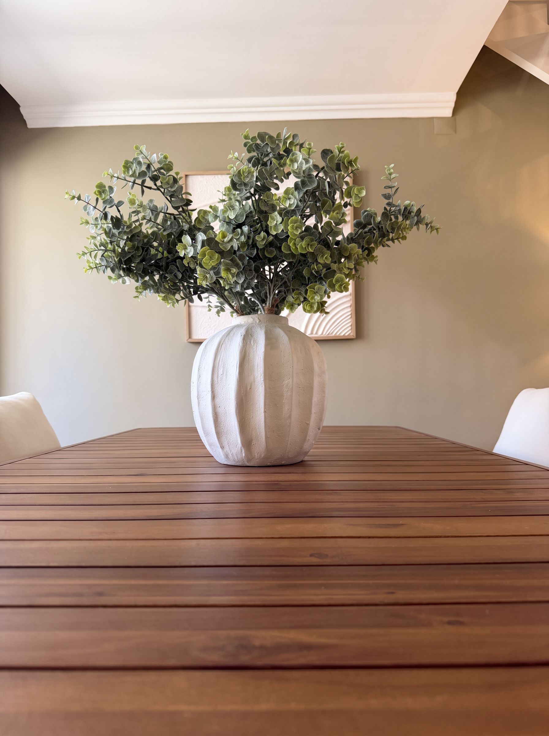 A wooden table with a large white vase containing green foliage.