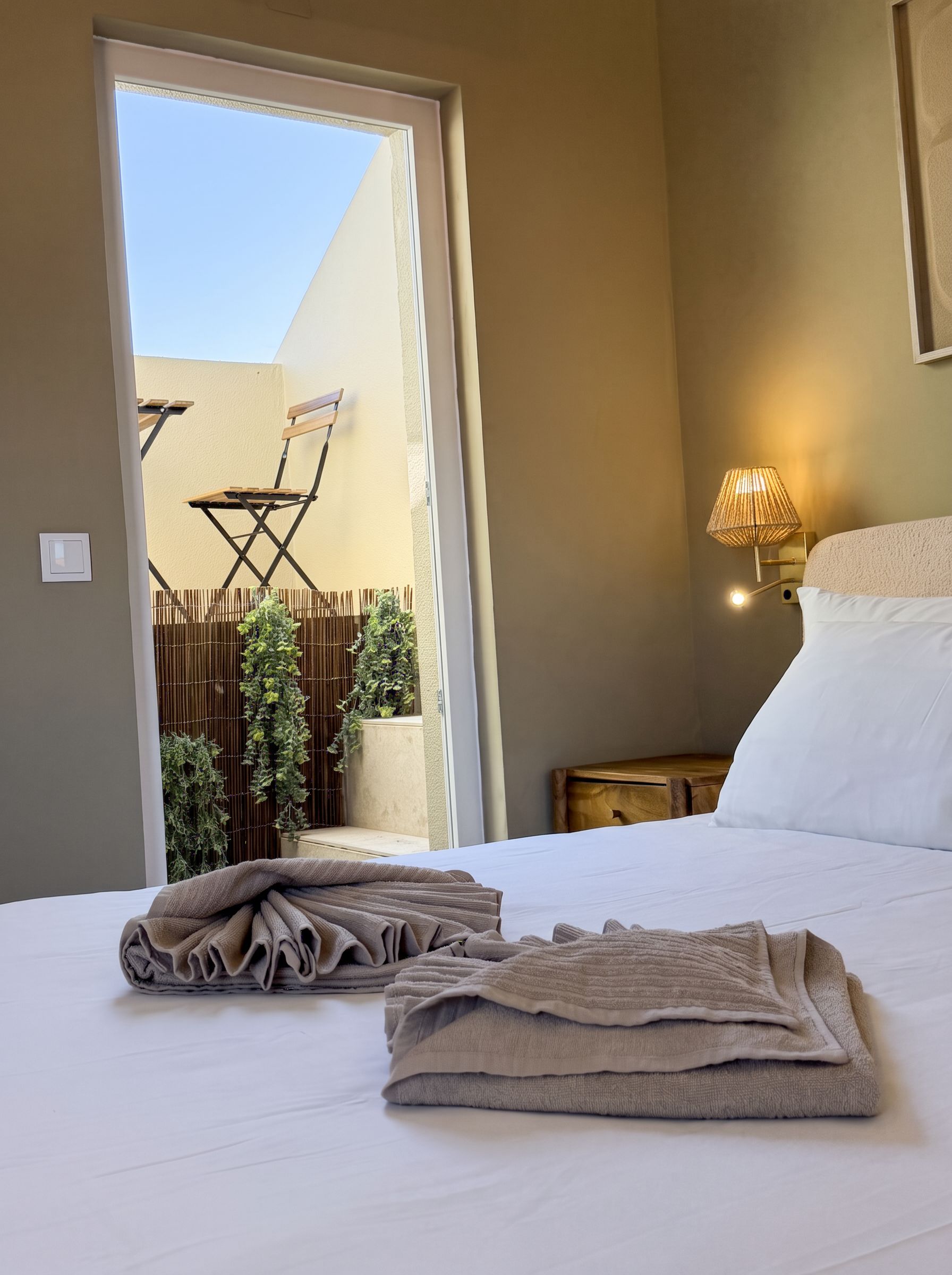 A hotel room with a bed, a nightstand, a lamp, a chair outside the door, and a view of the outside.
