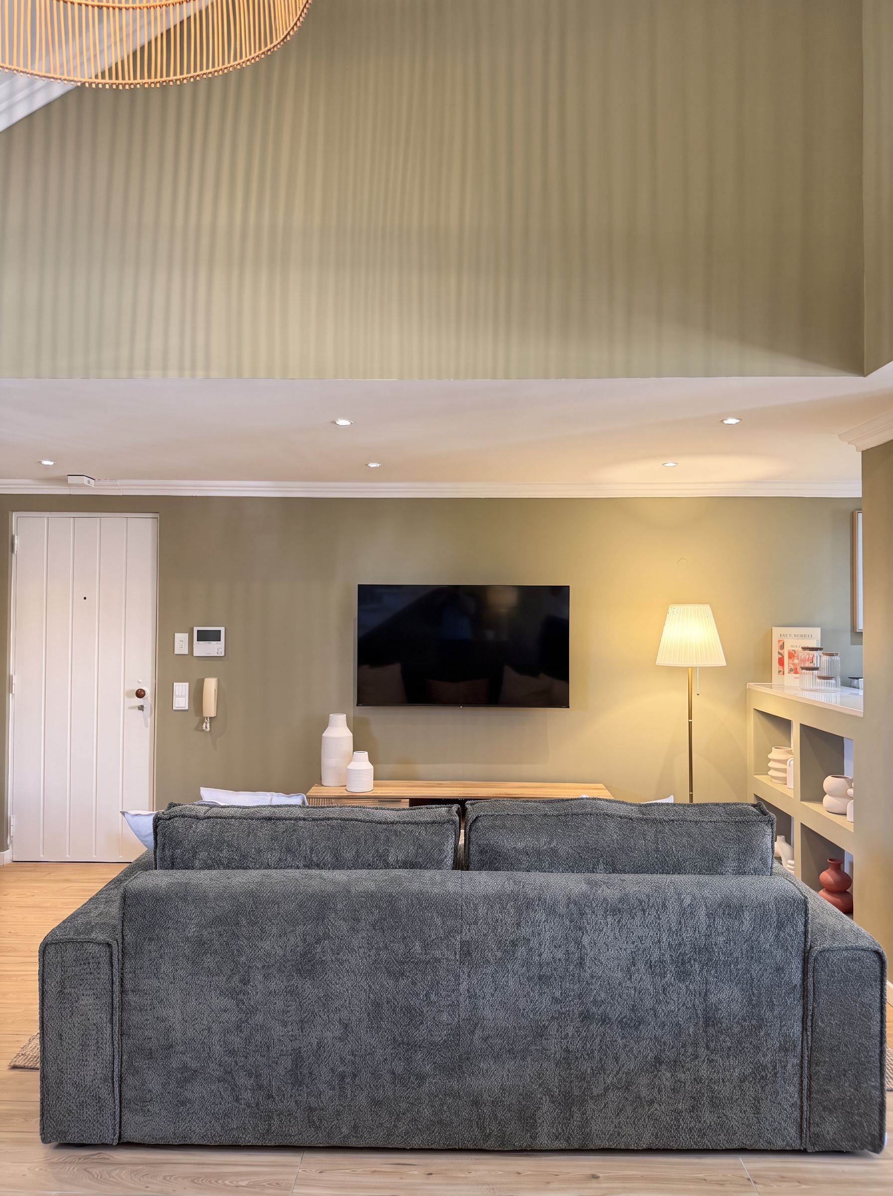 Un salon moderne avec un grand canapé d’angle gris, un téléviseur à écran plat fixé au mur, une table basse en bois et un lampadaire.