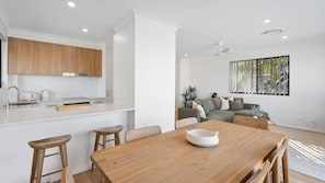 Eine moderne Küche mit weißen Schränken, einem Holztisch und einer Sitzecke inklusive Sofa und einem Fenster mit Jalousien.