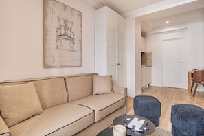 A modern living room with a beige sofa, a coffee table, and a blue ottoman. There is a painting on the wall and a door leading to a kitchen area. - Lovely apartment with AC - Center of Paris (Paris)