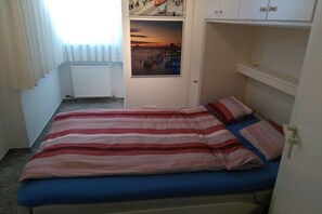 Une chambre à coucher avec un lit, une fenêtre avec des rideaux, un radiateur, une porte ornée d’une photographie représentant un paysage urbain et une armoire blanche.