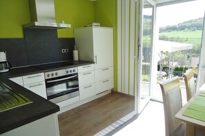 Une cuisine équipée d’une cuisinière, d’un four et de placards, un coin repas avec une table et des chaises, et une vue sur un paysage verdoyant par une porte vitrée.
