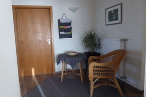 Une pièce avec une porte en bois, une petite table recouverte d’une nappe sombre, une chaise en osier, une plante en pot et un tableau encadré accroché au mur.