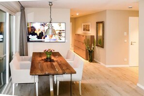 Ein Essbereich mit Holztisch und weißen Stühlen, einem an der Wand befestigten Fernseher und einer Tür zu einem anderen Raum.