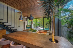 Ein modernes Interieur mit einem Holzesstisch, einem Fernseher, der 'NETFLIX' anzeigt, und Blick auf einen Swimmingpool und Grünflächen.