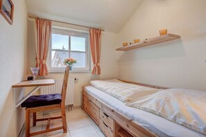 A small, compact bedroom with a single bed, a desk, and a chair. There is a window with curtains, a radiator, and a shelf with decorative items. - Holiday home with 3 bedrooms (Butjadingen)