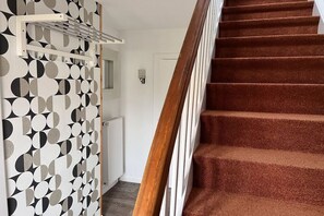 Un escalier recouvert d’une moquette marron et doté d’une rampe blanche, menant à une pièce dont un mur est orné d’un motif géométrique et qui contient un radiateur blanc.