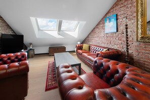 Ein Loft-Wohnzimmer mit hohen Decken, freiliegenden Backsteinwänden, einem großen Fenster, einem Ledersofa und einem Fernseher.
