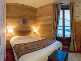 Quarto de cabana de madeira com uma cama, duas luminárias fixadas na parede, uma janela com cortinas e vista para o exterior.