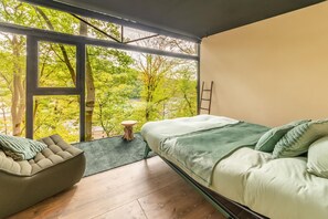 A bedroom with a bed, a chair, and a view of trees through large windows. - The Serene Escape – Olivenise Cabin (La Roche-en-Ardenne)
