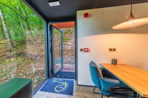 A modern interior with a wooden dining table, blue chair, and a view of a forest through a glass door. - The Serene Escape – Olivenise Cabin (La Roche-en-Ardenne)