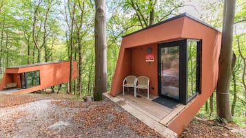 Uma cabana pequena e moderna com porta de vidro, duas cadeiras e um deck de madeira numa floresta.