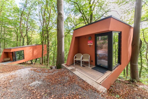 Una petita cabana moderna amb una porta de vidre, dues cadires i un balcó de fusta al bosc.