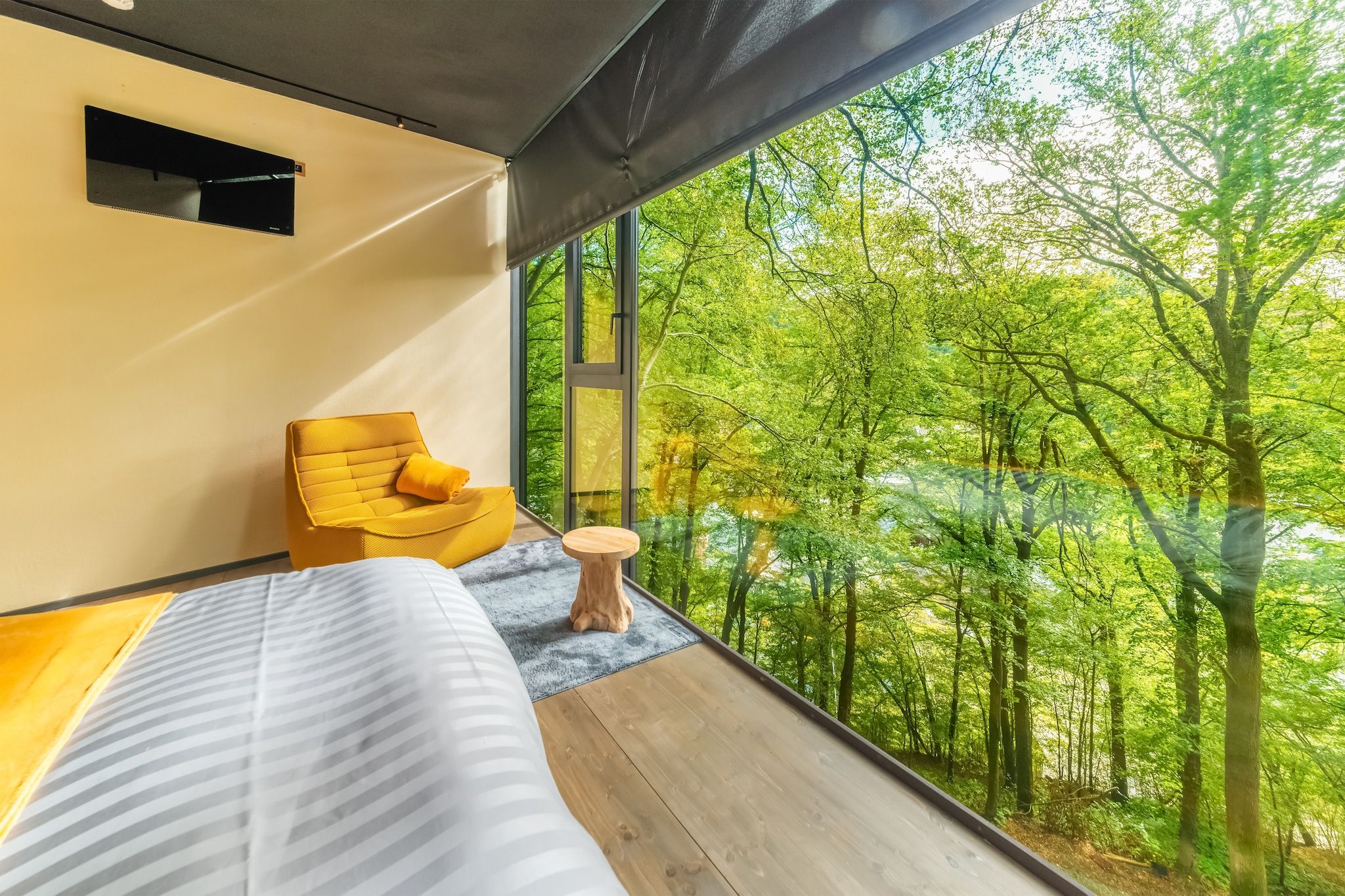 A bedroom with a large window, a bed, a yellow chair, and a small wooden table.