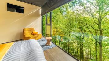A bedroom with a large window, a bed, a yellow chair, and a small wooden table.