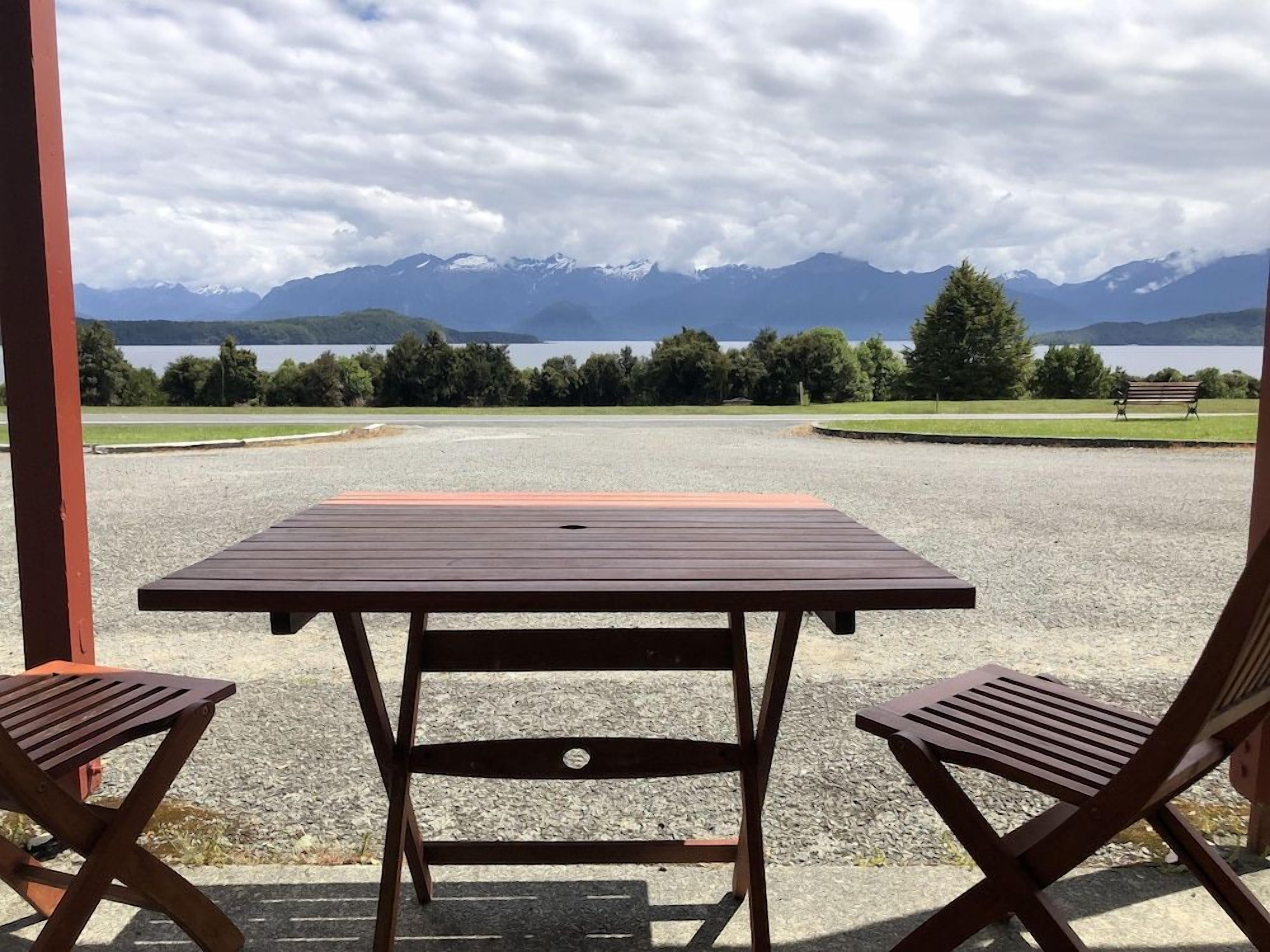 A covered outdoor seating area with a table and two chairs, overlooking a lake and mountains.