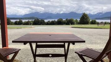 A covered outdoor seating area with a table and two chairs, overlooking a lake and mountains.