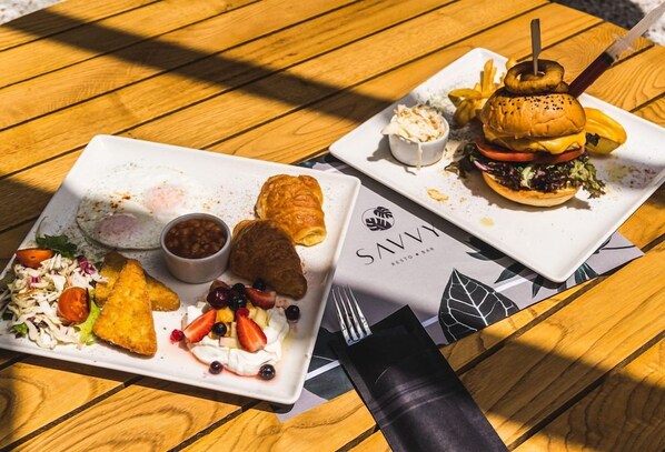 Two plates of food on a wooden table, one with a croissant, beans, and salad, and the other with a burger, fries, and coleslaw. - Canadian Annex (Zakynthos)