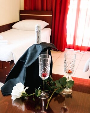 Habitación de hotel con una cama bien tendida, una botella de champán, dos copas de cristal y una flor blanca sobre la mesa.