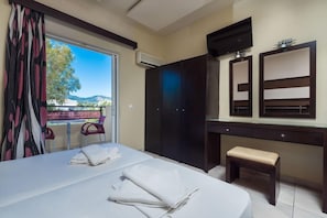 A hotel room with a bed, a desk, a bench, a wardrobe, and a large window with a view of the outdoors. - Canadian Annex (Zakynthos)