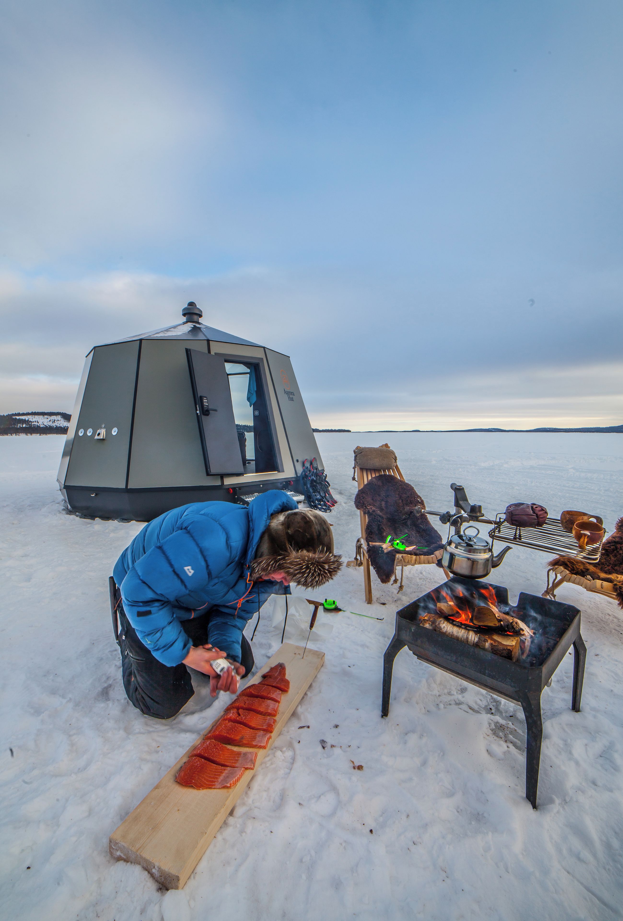 En person som lagar mat utomhus med hjälp av ett portabelt stormkök, ett tält och en pulka i ett snötäckt landskap.