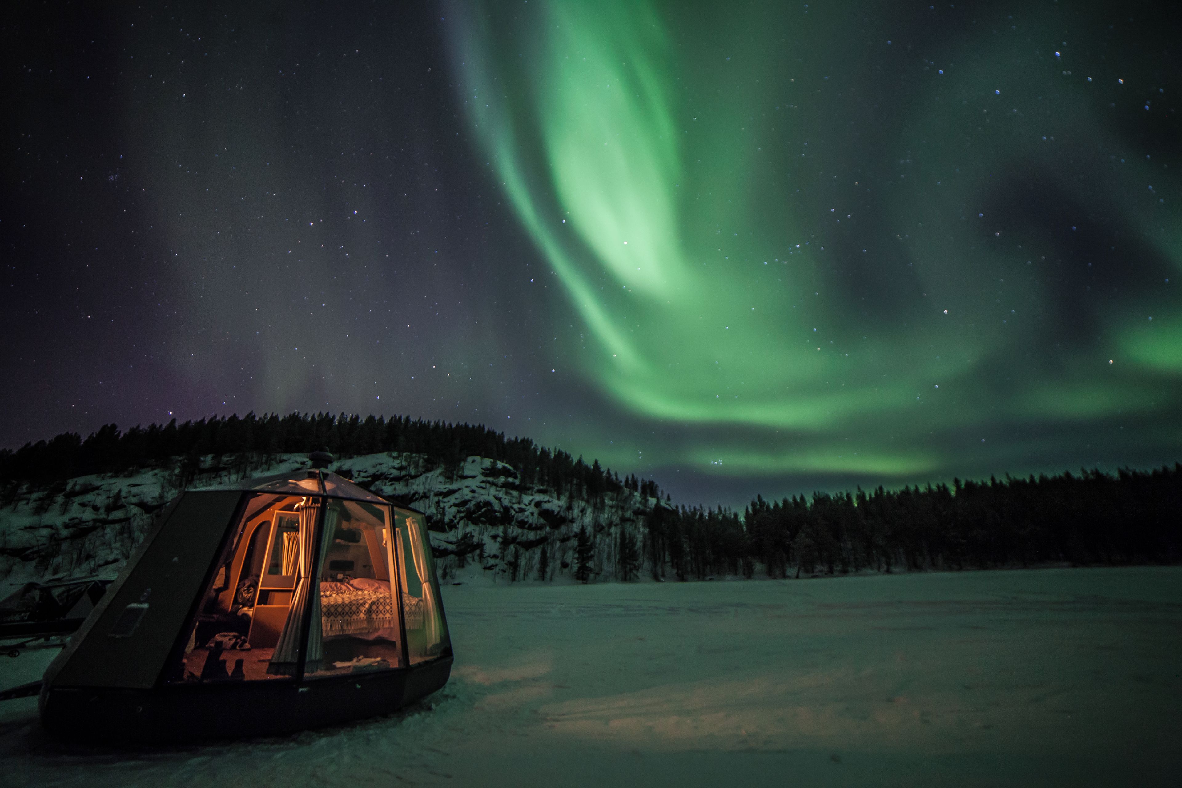 Ett snötäckt landskap med en tältliknande konstruktion och utsikt över norrskenet.