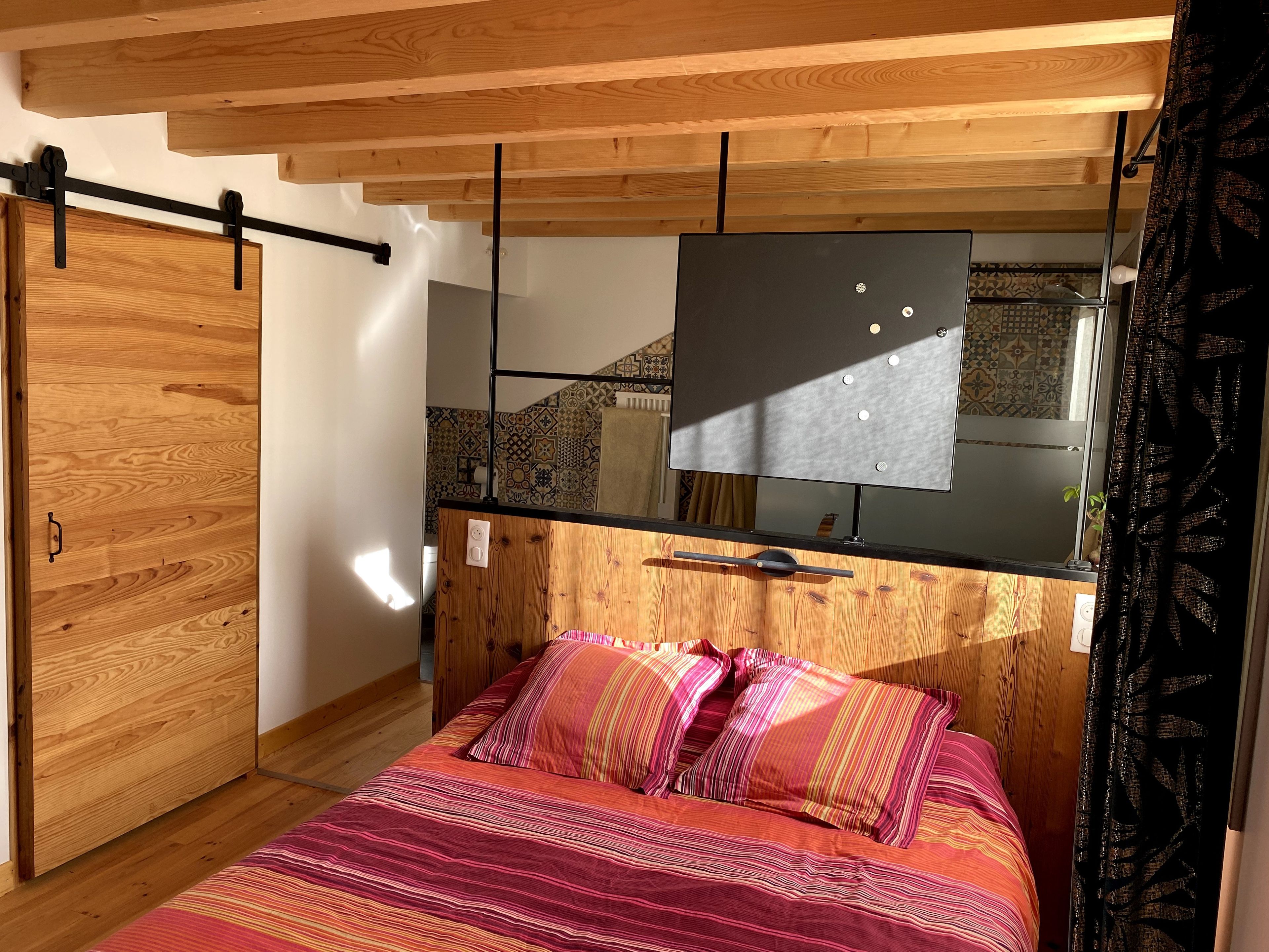 Une chambre avec un plafond en bois, un lit avec une literie à rayures, un téléviseur noir posé sur un meuble en bois et un mur à motifs.