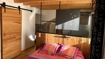 Une chambre avec un plafond en bois, un lit avec une literie à rayures, un téléviseur noir posé sur un meuble en bois et un mur à motifs.