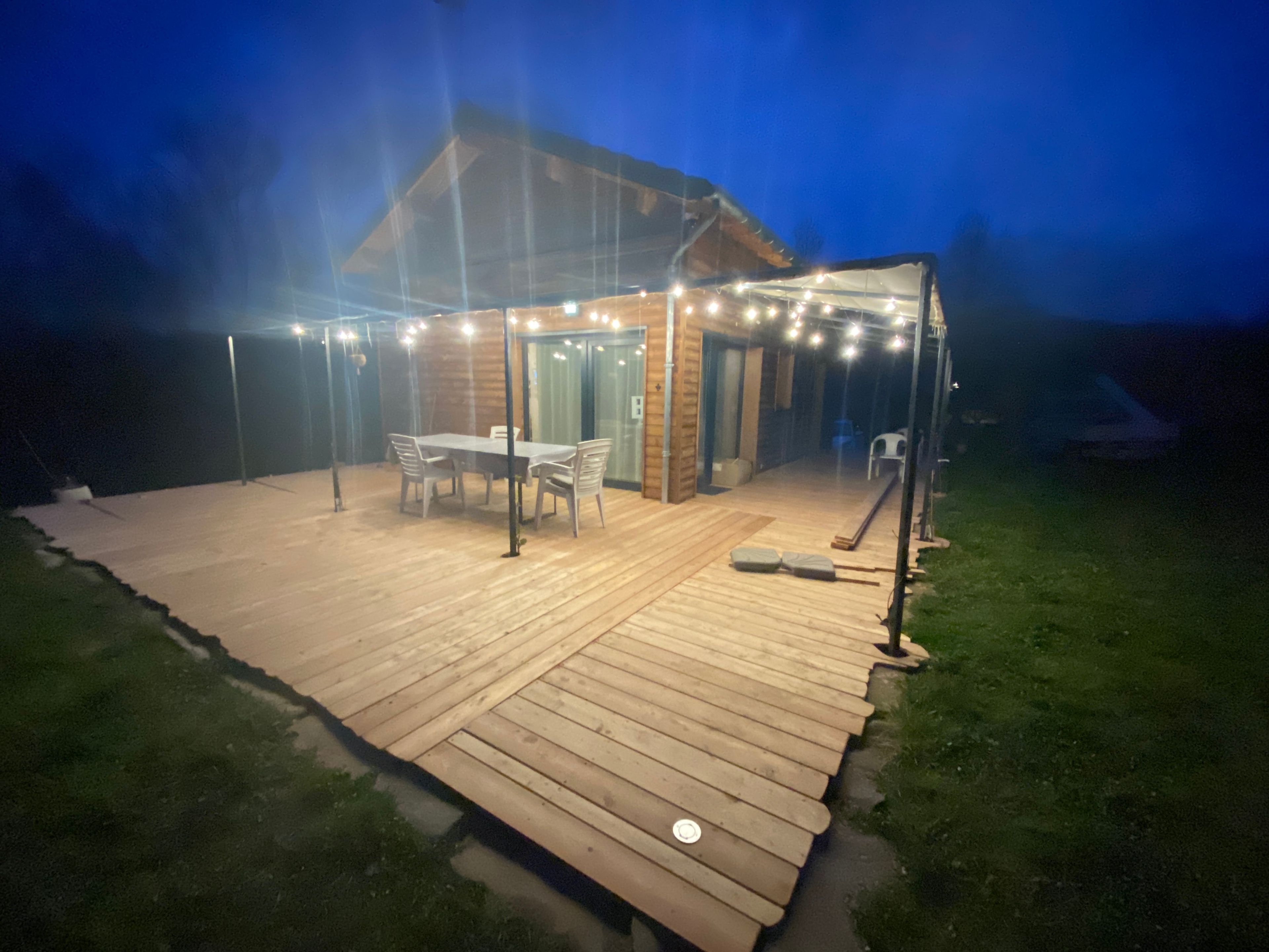 Una terraza de madera con una mesa y sillas, rodeada por una zona cubierta con luces colgantes.