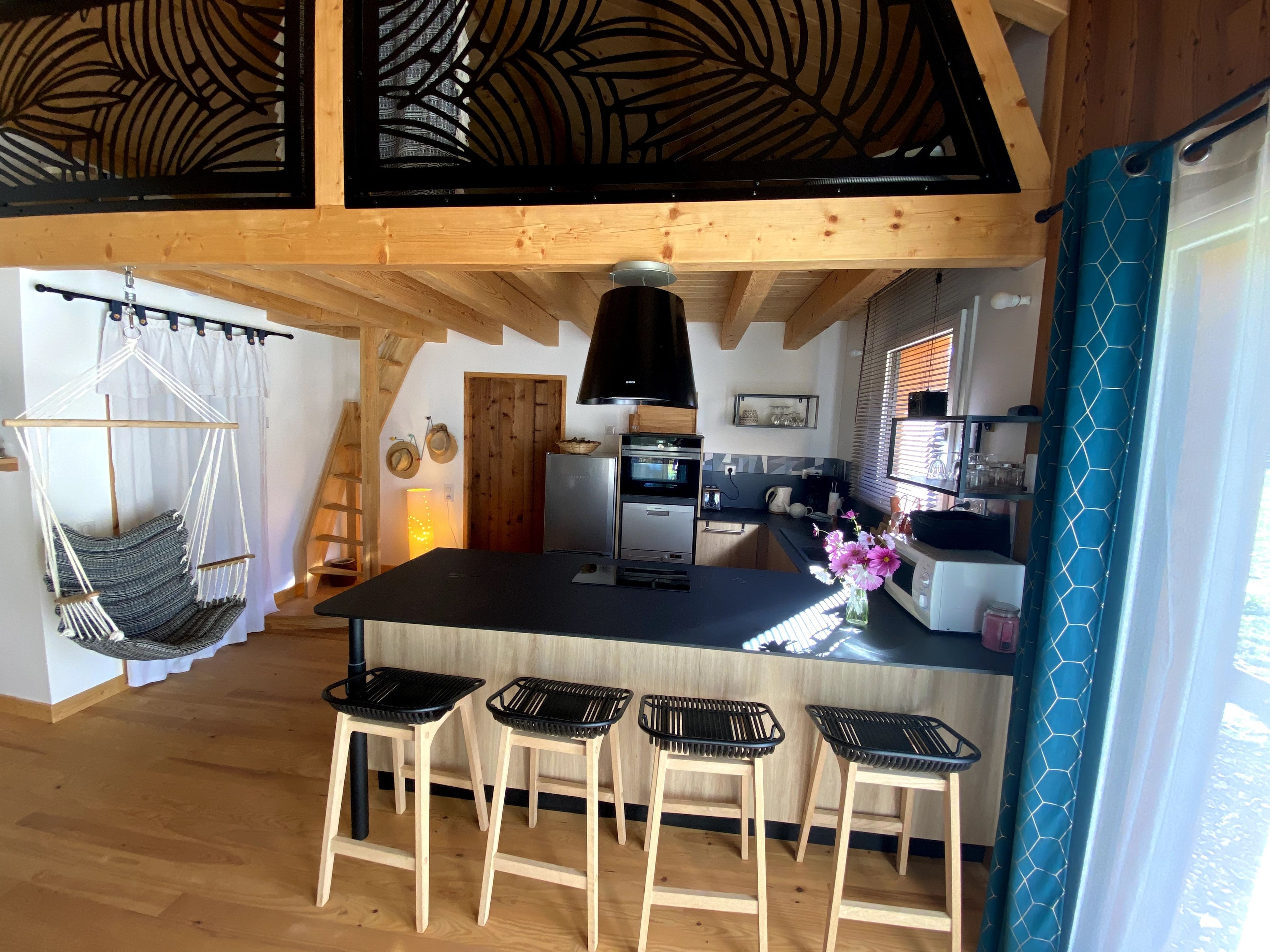 Una cocina con una isla central, taburetes de madera, una silla colgante y una ventana con cortinas.