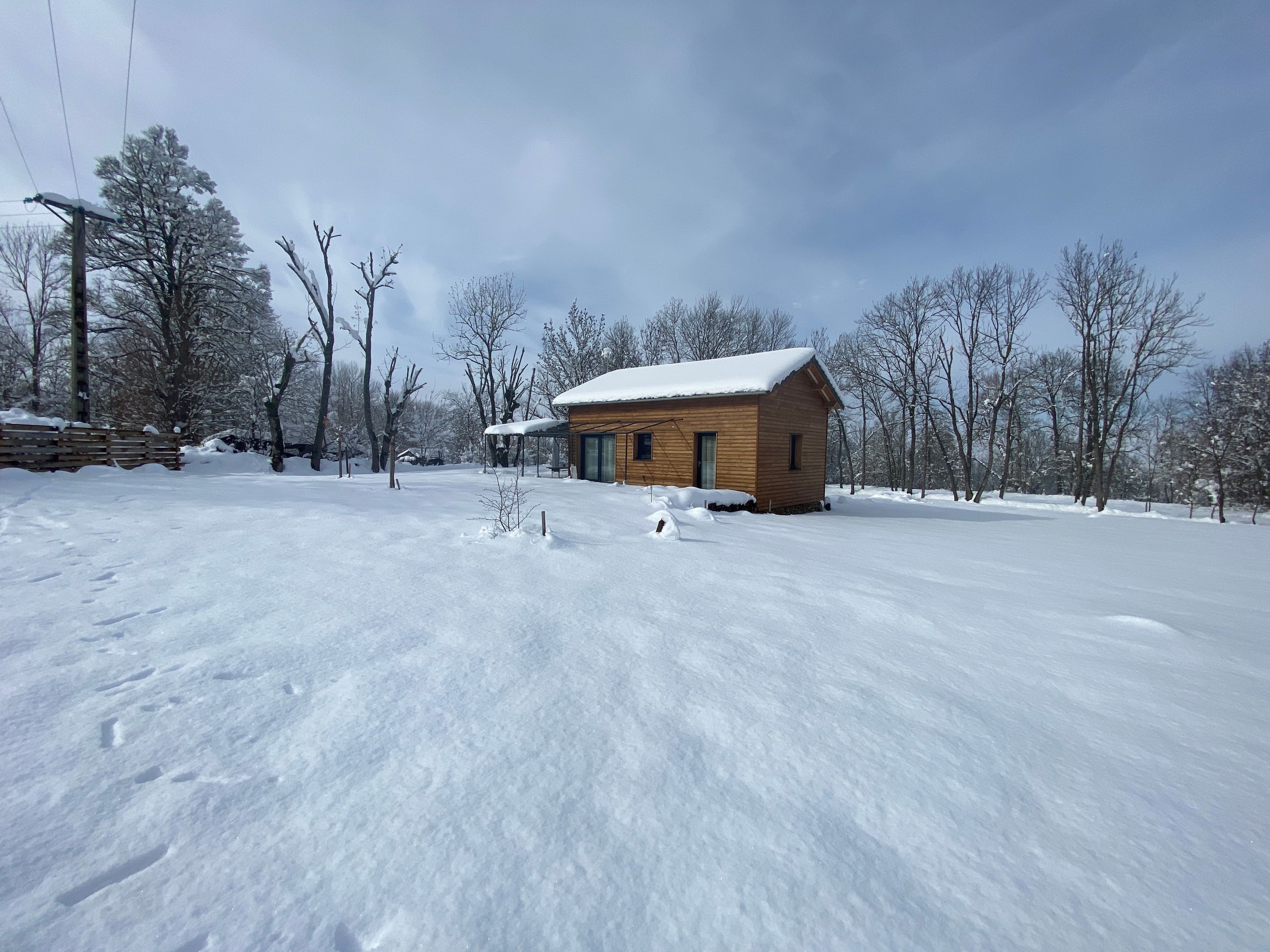 Une cabane en bois dont le toit est recouvert de neige, entourée d’un paysage enneigé et d’arbres sans feuilles.